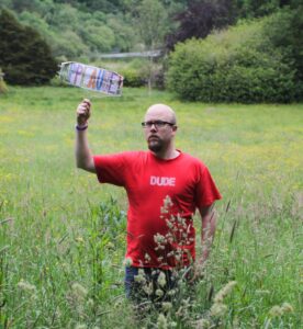 Kalle Ryan in a large green field. Red t-shirt with white lettering tht says "Dude". Holding up a handmade sign that says "Applause"