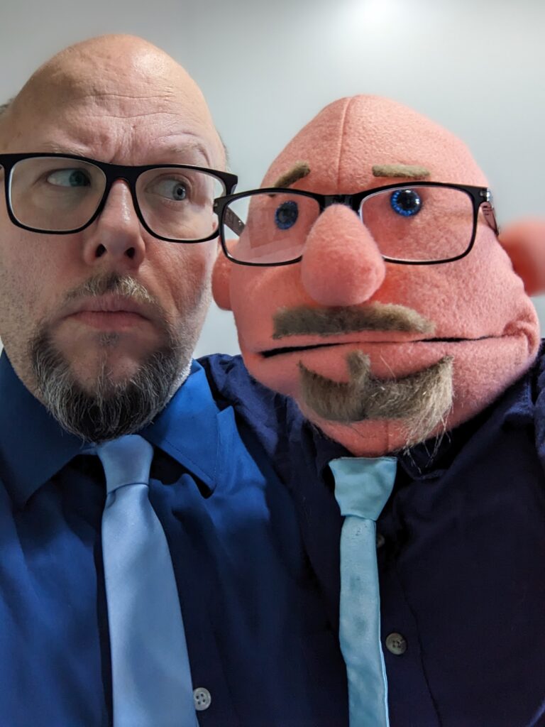 Kalle Ryan , navy shirt, blue tie, sideyeing his Kalle Ryan puppet/muppet who is wearing the same clothes